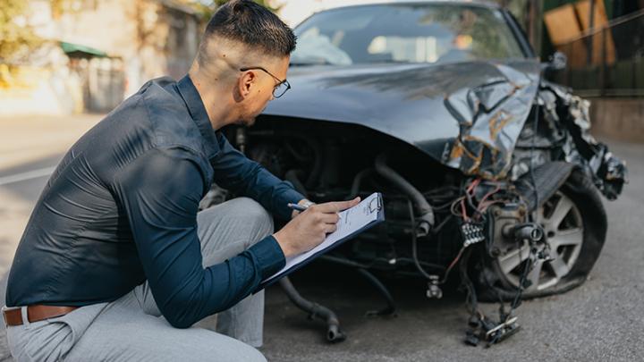 A young male claims adjuster with a clipboard crouches in front of a black damaged vehicle 