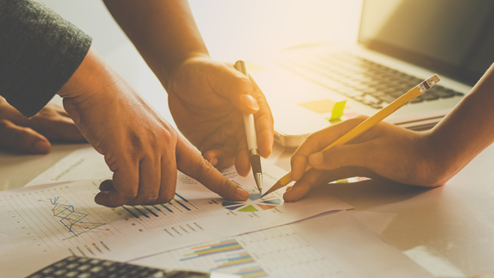 Three hands pointing down on a paper with a pie graph in the middle 