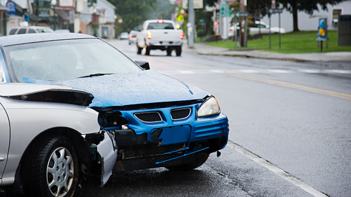 A blue car and a silver car have crashed on a rainy street. The hoods and bumpers of both cars are damaged. 