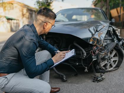 A young male claims adjuster with a clipboard crouches in front of a black damaged vehicle 