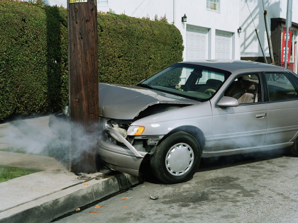 A silver sedan has crashed into a telephone pole. The hood of the car is crumpled and smoke is coming from the engine. 