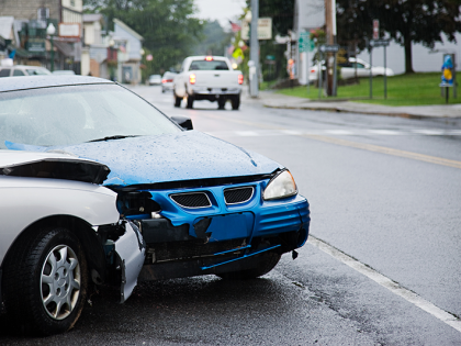 A blue car and a silver car have crashed on a rainy street. The hoods and bumpers of both cars are damaged. 