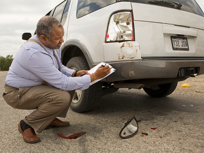 A claims adjuster crouches with a clipboard at the rear of a white mini-van assessing damage to the rear tail light and bumper. 